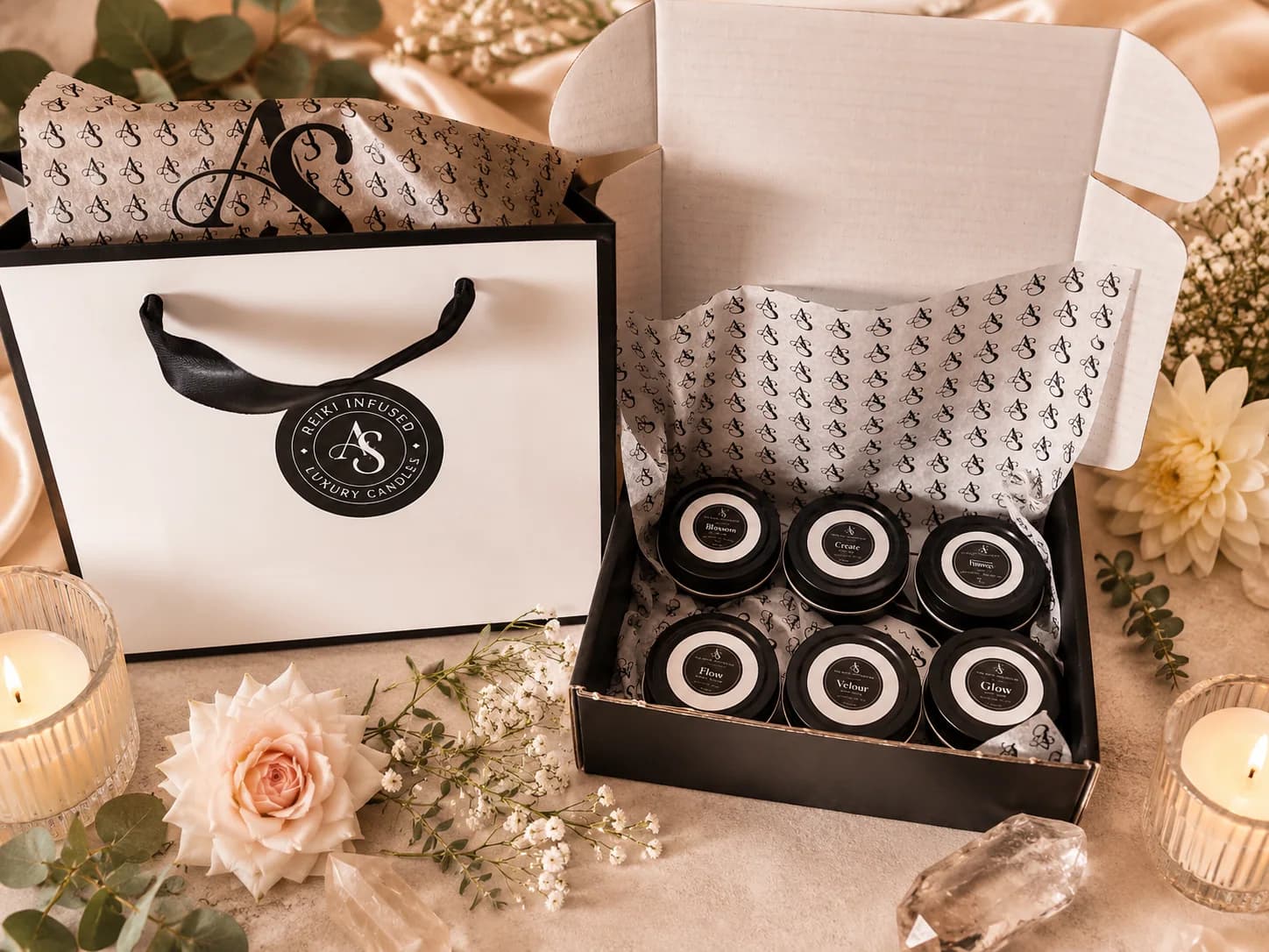 Six mini candles in an open gift box beside a branded bag, tissue and flowers